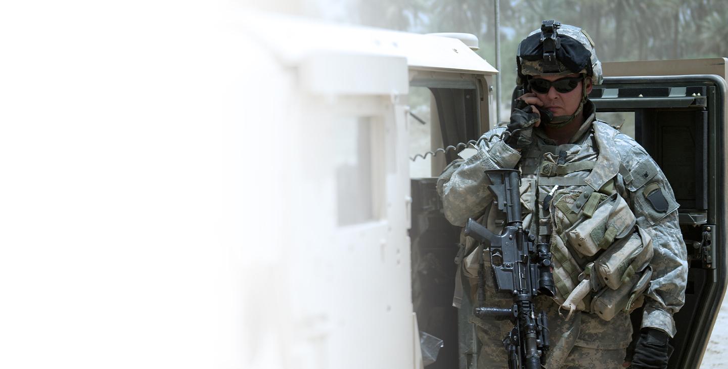 一名男子身穿战斗装备，手持军用步枪，对着从一辆悍马延伸出来的收音机讲话